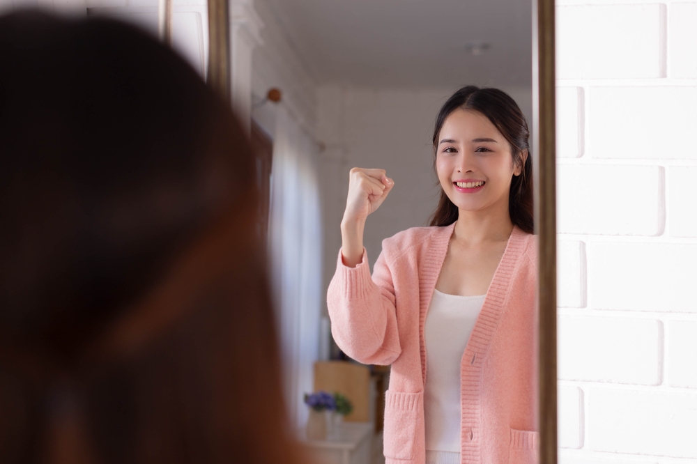 Smiling,Beautiful,Asian,Woman,Looking,At,Herself,In,The,Bathroom Smiling woman looking in the mirror and raising her fist in confidence, symbolising self-reflection and personal growth inspired by BaZi analysis in Singapore